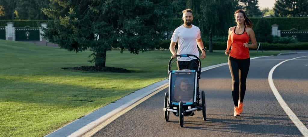 Ieși la alergare cu stil – căruciorul sport și accesoriile care fac diferența Părinți alergând cu un cărucior sport dotat cu husă de protecție și spațiu de depozitare, într-un parc verde.