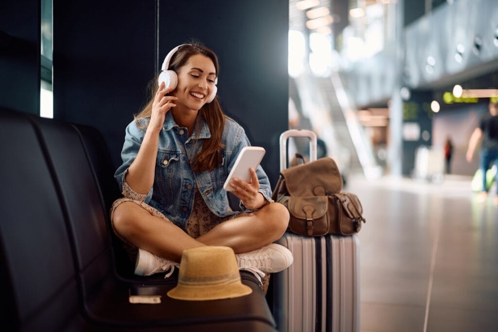 Cum să nu rămâi fără baterie în aeroport Femeie relaxată pe bancheta unui aeroport, uitându-se la telefon, exemplificând consumul bateriei din cauza ecranului