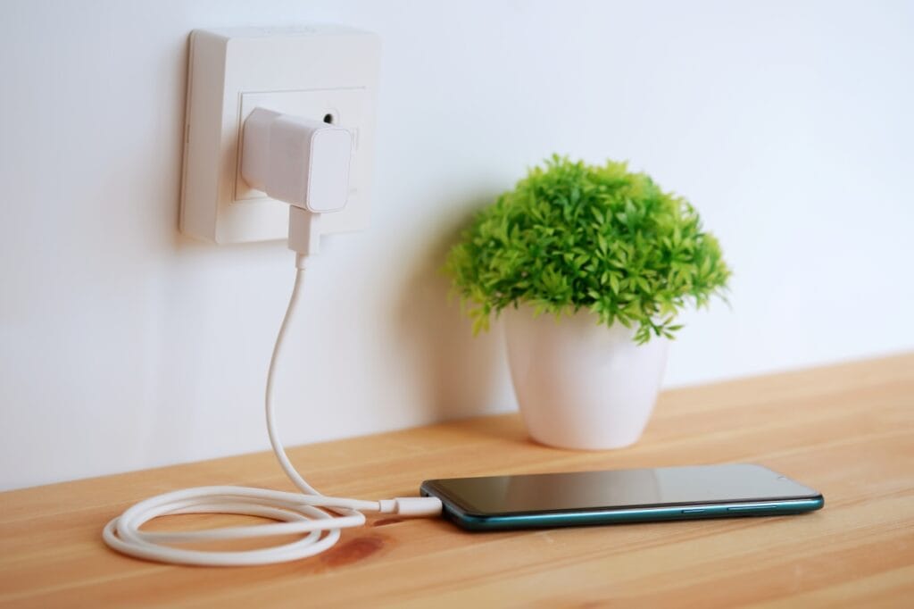 Ce poți face ca bateria telefonului să țină mai mult Smartphone în încărcare cu cablu alb conectat la priză, pe un birou din lemn, alături de o plantă decorativă.