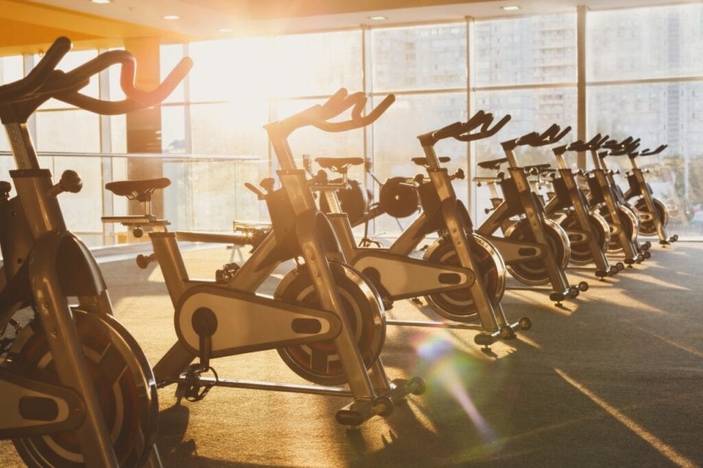 Întreținerea corectă a bicicletelor de fitness: secretul longevității Biciclete de fitness indoor aliniate într-o sală de sport iluminată de soare, întreținute și curate.