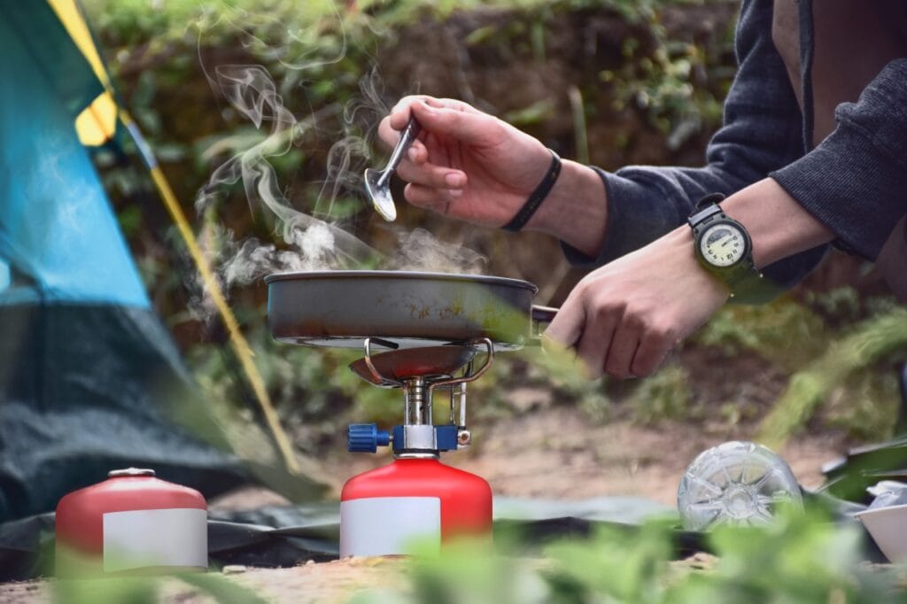 Gătitul în siguranță în camping – cum folosim corect un arzător portabil Persoană gătește în aer liber cu un arzător de camping portabil, pe gaz, lângă un cort