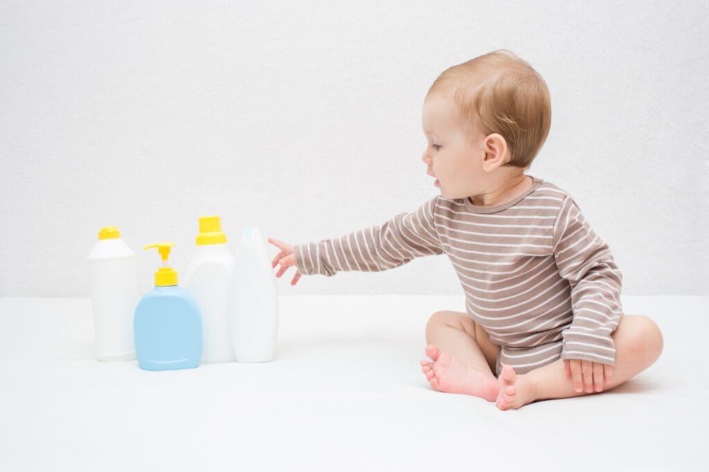 Ce șampon alegi pentru copil? Contează tipul de păr! Copil mic în pijamale lunguțe atinge sticle de șampon, simbolizând alegerea corectă pentru părul sensibil al bebelușului