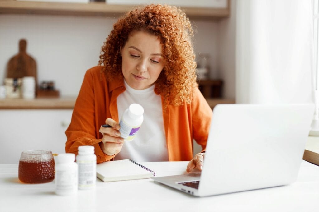 Ce nu știi despre suplimente poate afecta tratamentul tău Persoană analizând compoziția unui supliment alimentar lângă un laptop, simbolizând nevoia de informare în contextul terapiei medicamentoase.