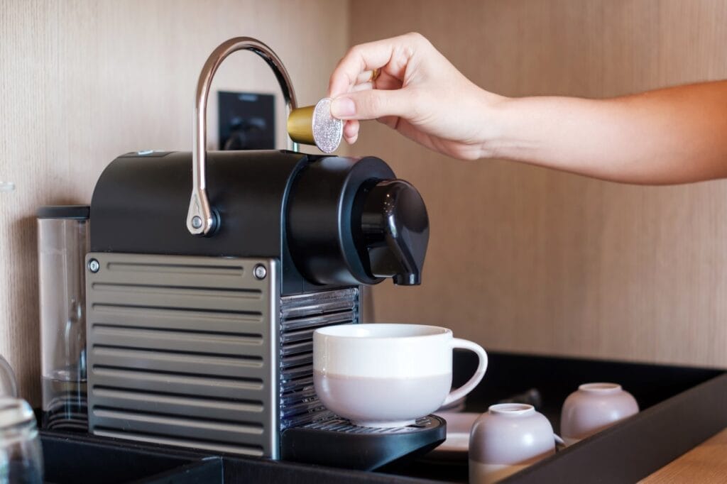 Cafea instant la standarde înalte – espressor cu capsule în acțiune Mână care introduce o capsulă într-un espressor automat de cafea, cu o ceașcă pregătită.