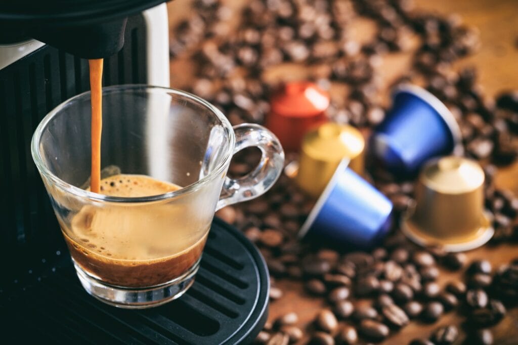 Gust instant, zero compromisuri – espressorul cu capsule Cafea proaspătă turnându-se într-o ceașcă de la un espressor cu capsule, pe un fundal cu capsule colorate și boabe de cafea.