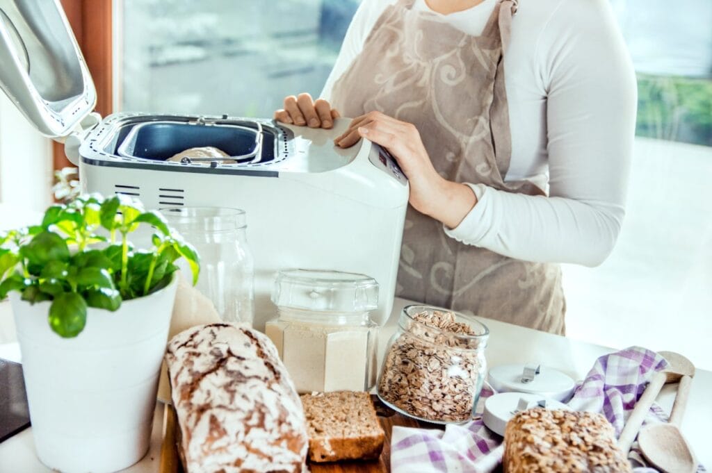Pâine de casă cu un consum redus de energie Persoană verificând mașina de pâine în bucătărie, înconjurată de pâine de casă și borcane cu ingrediente