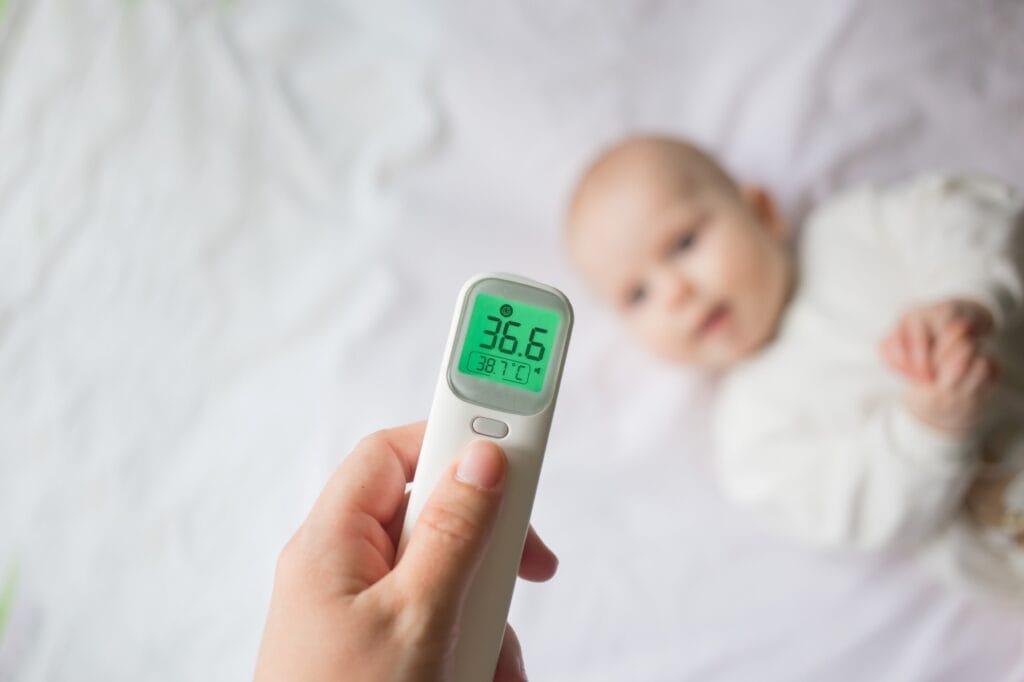 Este timpul să îți schimbi termometrul? Mână ținând termometru infraroșu digital în fața unui bebeluș, verificând temperatura