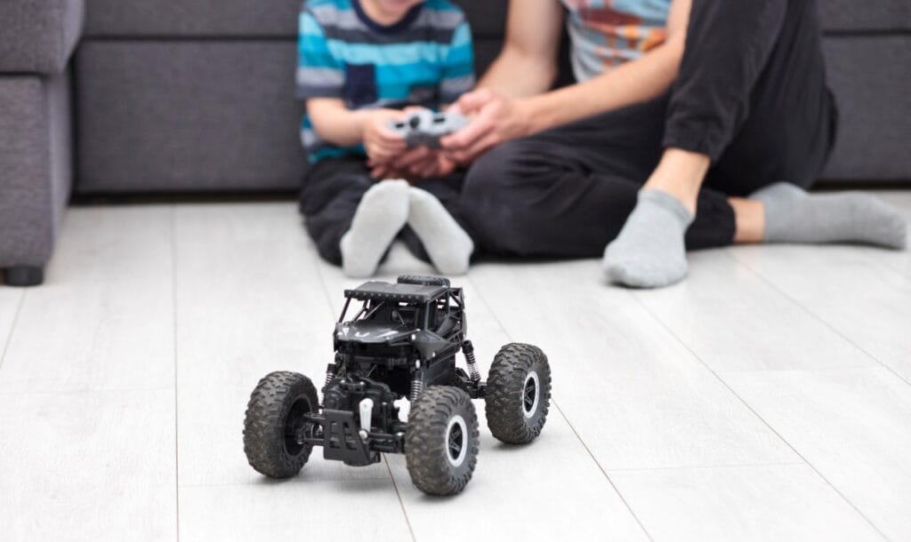 Cum arată joaca atunci când telecomenzile fac diferența Mașină cu telecomandă off-road în prim plan, copil și părinte controlează de pe canapea