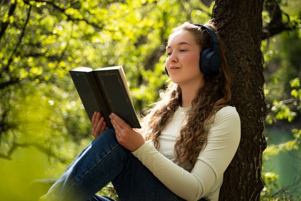 Ce să alegi: bestseller sau carte premiată pentru copii 12-14 ani? Adolescentă relaxată citind carte și ascultând muzică în parc, importanța alegerii corecte între bestseller și titlu premiat