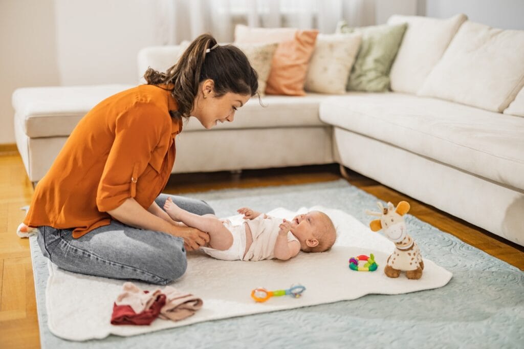 mamă schimbă scutecul bebelușului pe covor într-un living luminos, ghid frecvență schimbare scutec, îngrijire bebeluși acasă