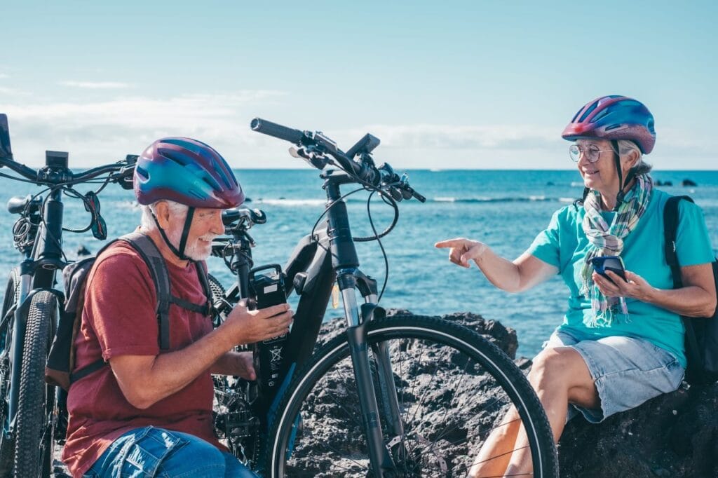 Seniori verificând aplicația smart a bicicletei electrice lângă mare, setare autonomie și funcții de control