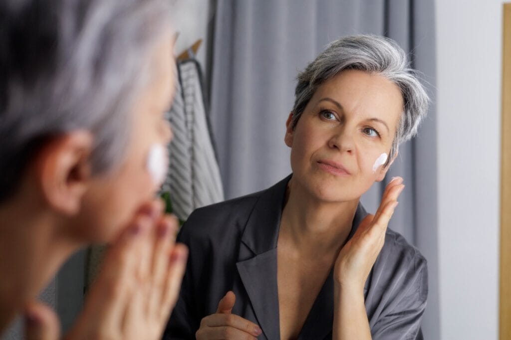 femeie matură aplică cremă antirid pe față în fața oglinzii, rutină de zi și de noapte