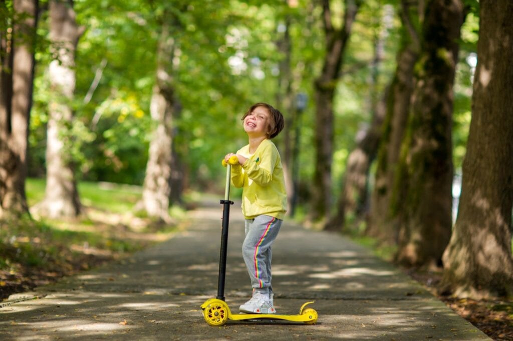 copil fericit poartă pantaloni de trening gri și hanorac galben pe trotinetă, joacă în parc